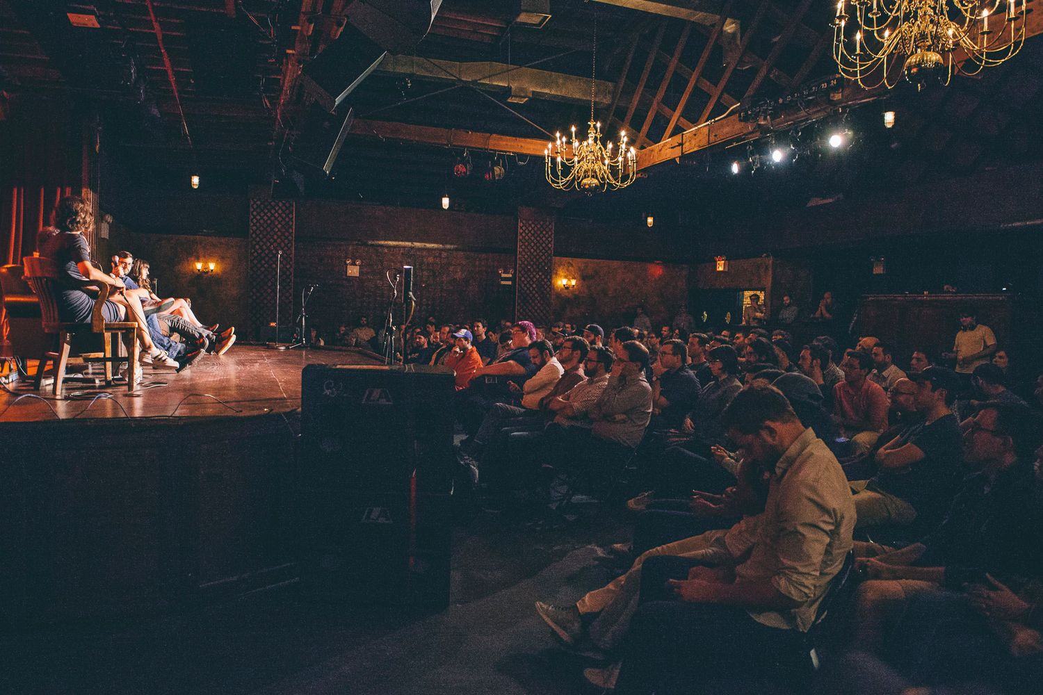 Audience seated in rustic venue watching intimate stage panel with chandelier lighting.