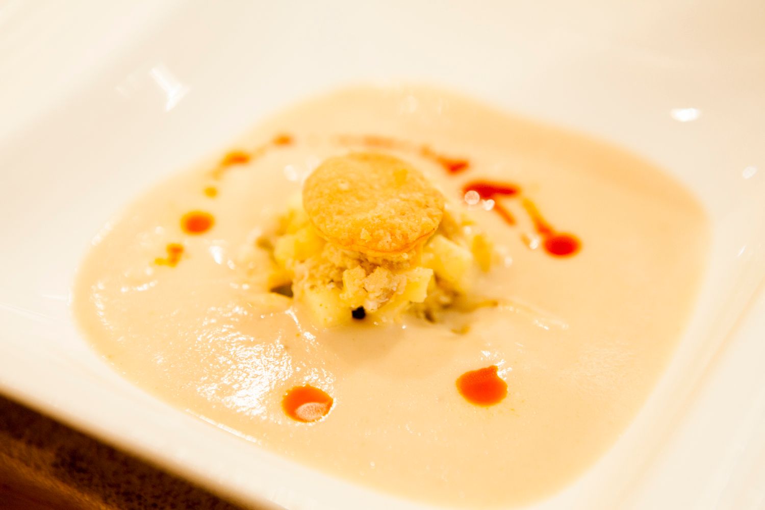 Elegant creamy soup with a golden fried scallop, garnished with red sauce drops on a white square plate.