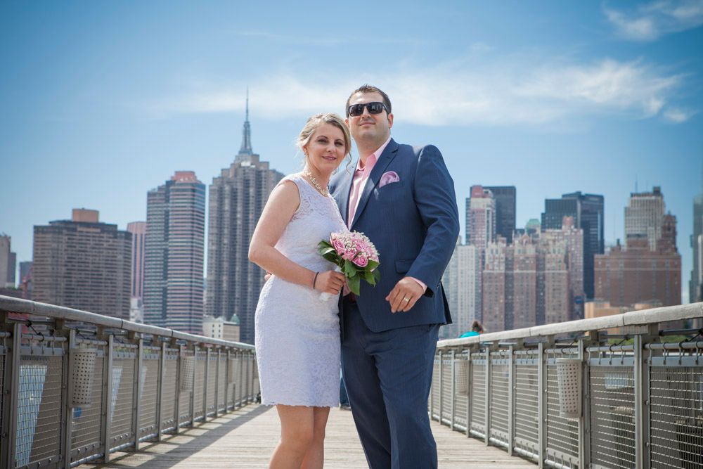 Wedding couple poses on NYC waterfront pier, groom in navy suit and bride in white dress with pink bouquet.