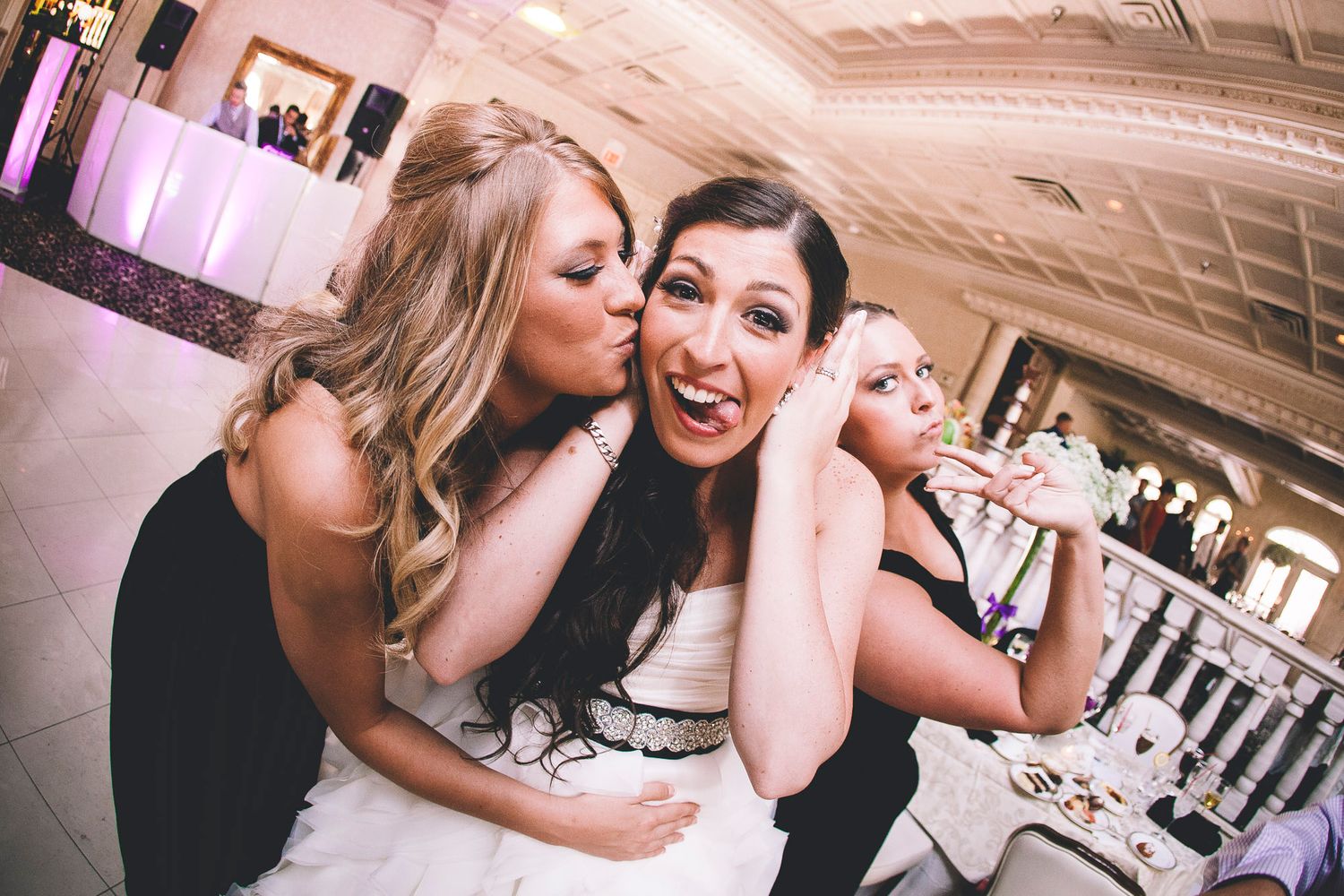 Bridesmaid kissing bride on the cheek while another guest laughs at the wedding reception party.