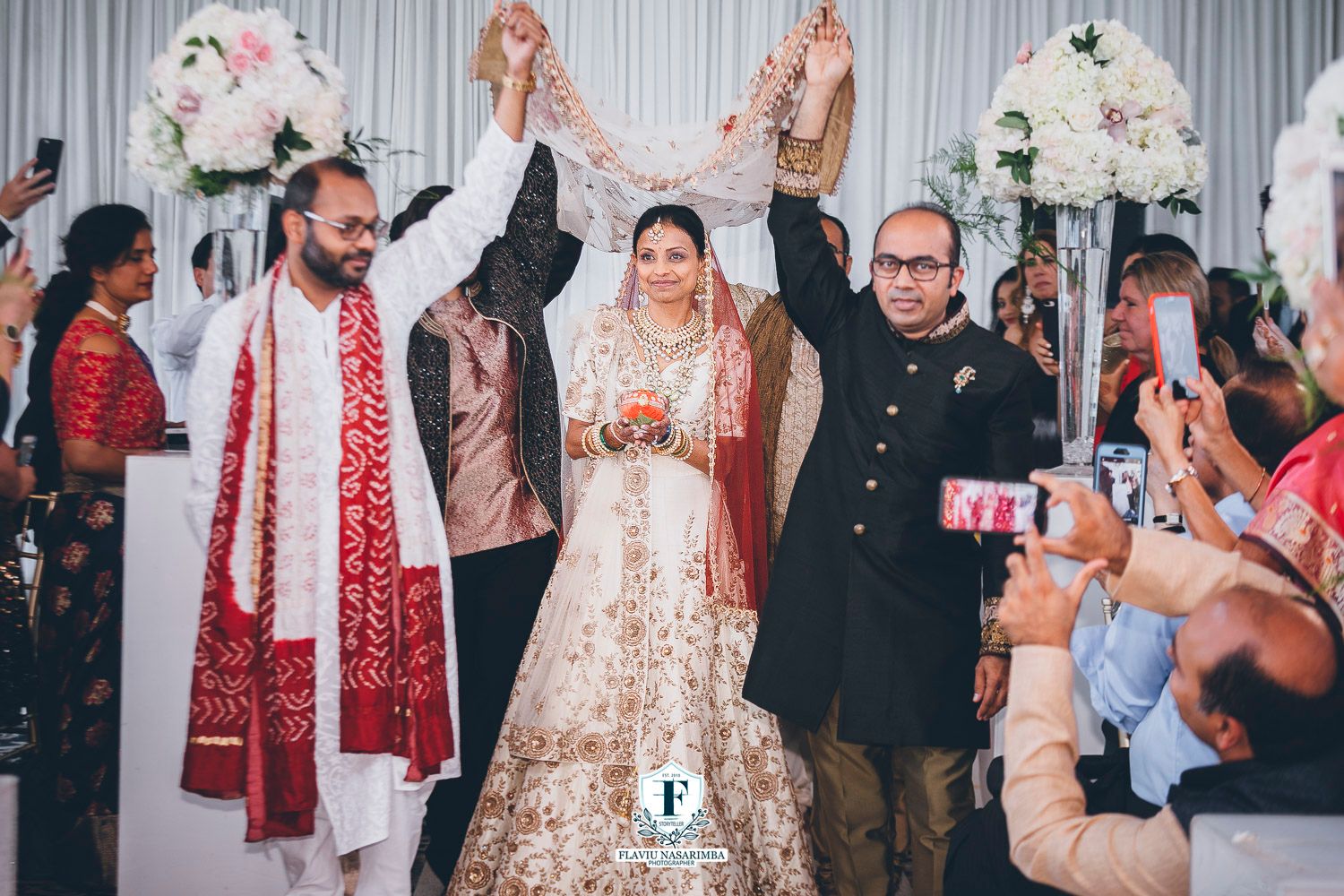 Bride in gold lehenga escorted by two men raising hands in celebration at Indian wedding entrance.