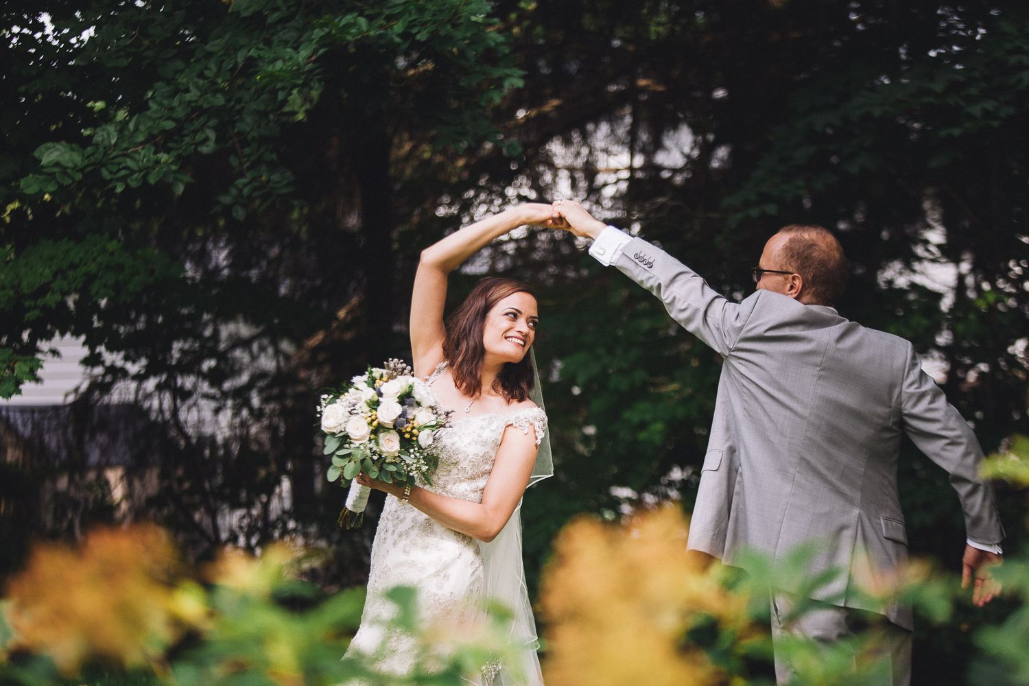 Groom twirling bride in a lace wedding gown among golden flowers in a lush garden setting.