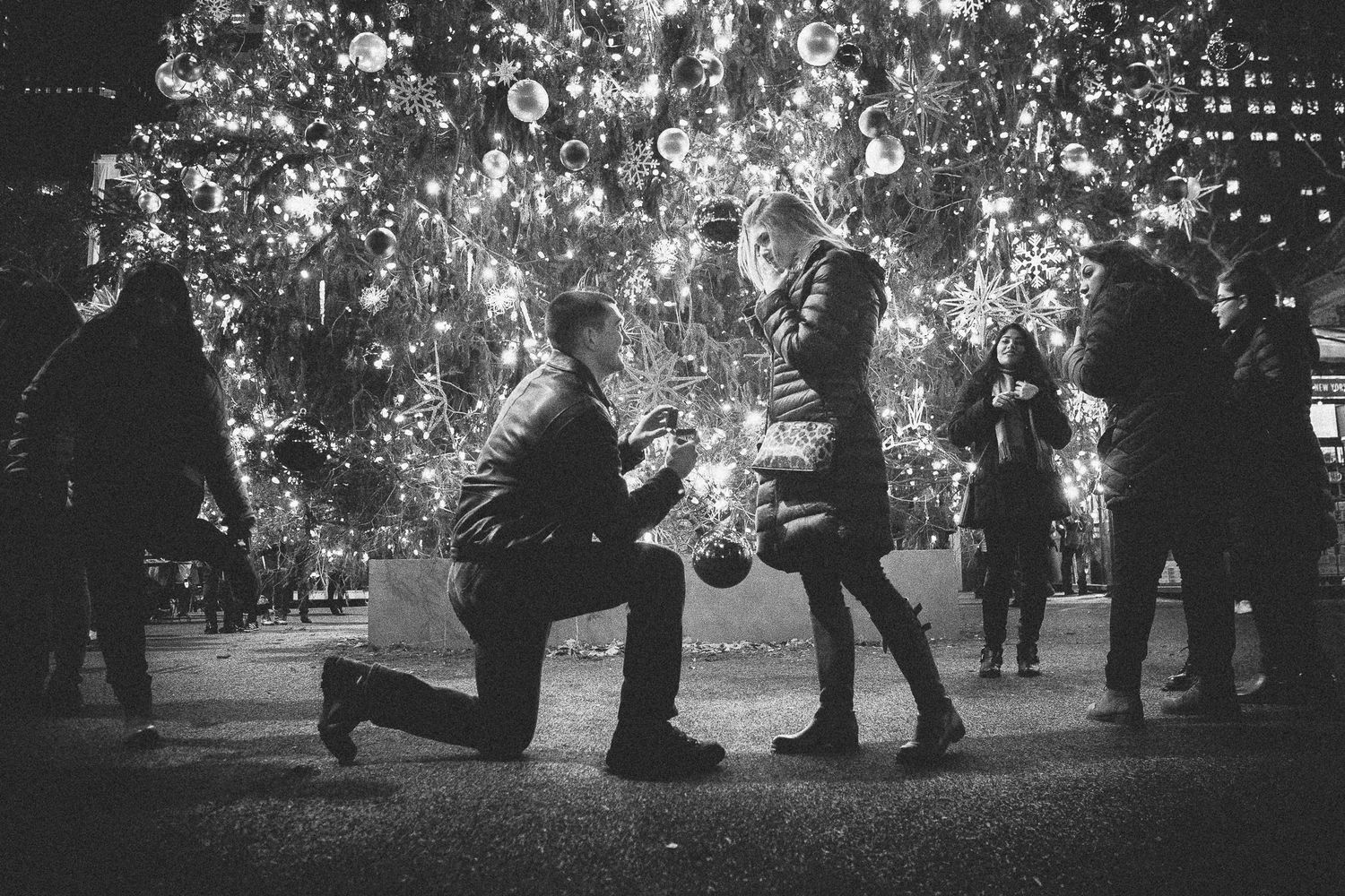Black and white photo of marriage proposal before glowing Christmas tree with onlookers nearby.