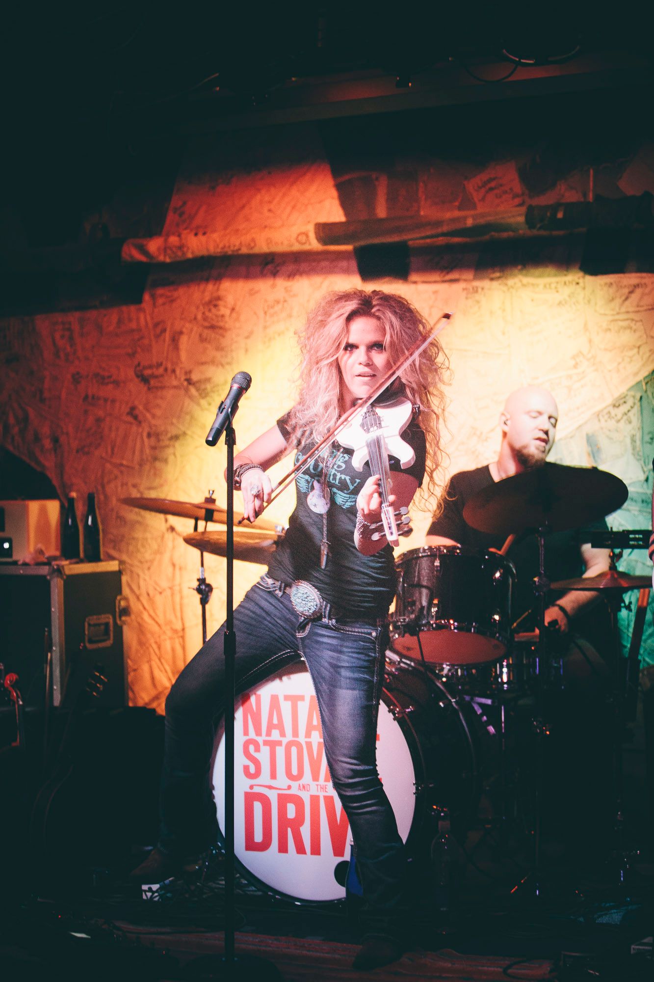 Female violinist performs on stage with drummer behind her, band name visible on bass drum.