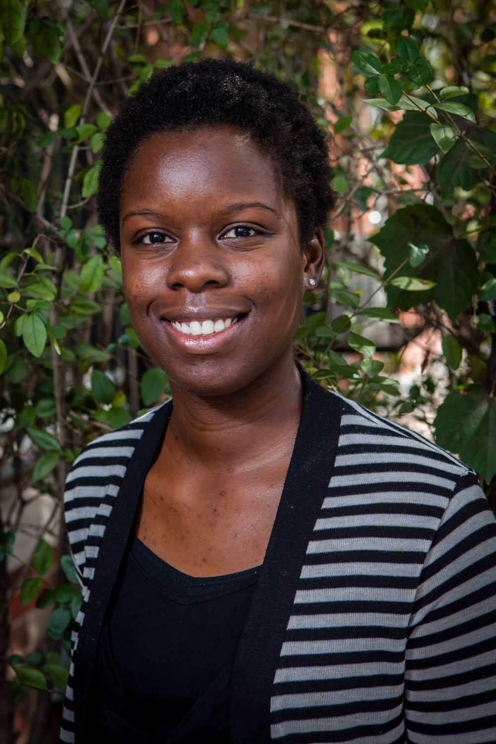 Smiling young woman in striped cardigan posing outdoors with green foliage background.