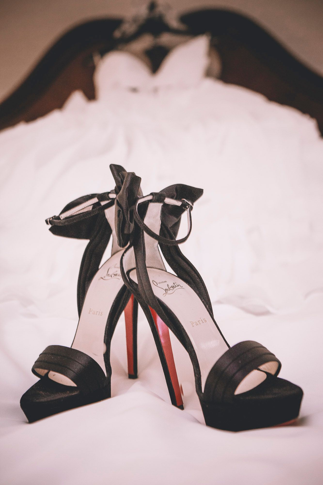 Black Christian Louboutin platform heels with red soles displayed on white bed with wedding dress behind.