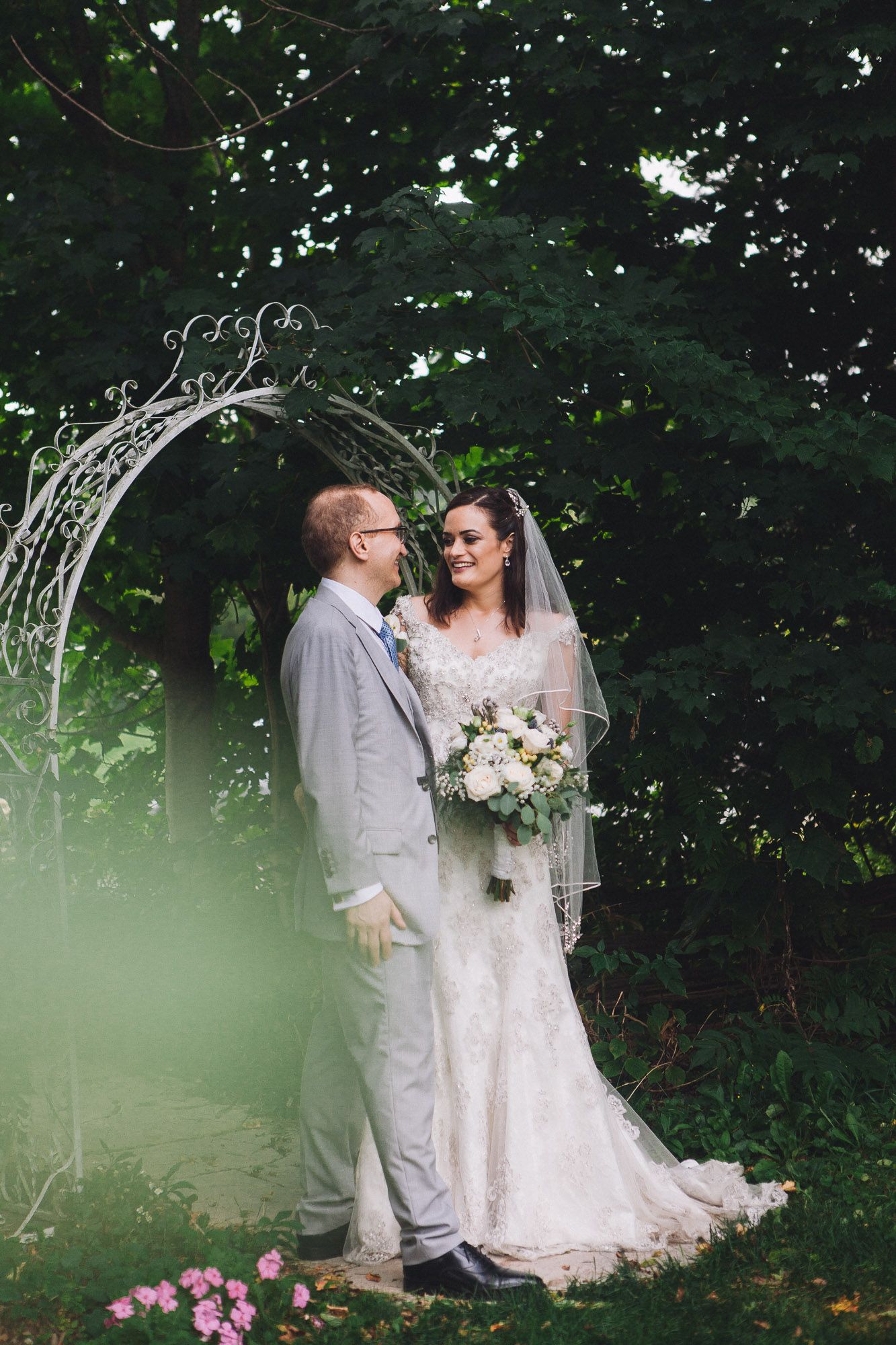 Bride and groom standing under a white iron arch surrounded by green trees.