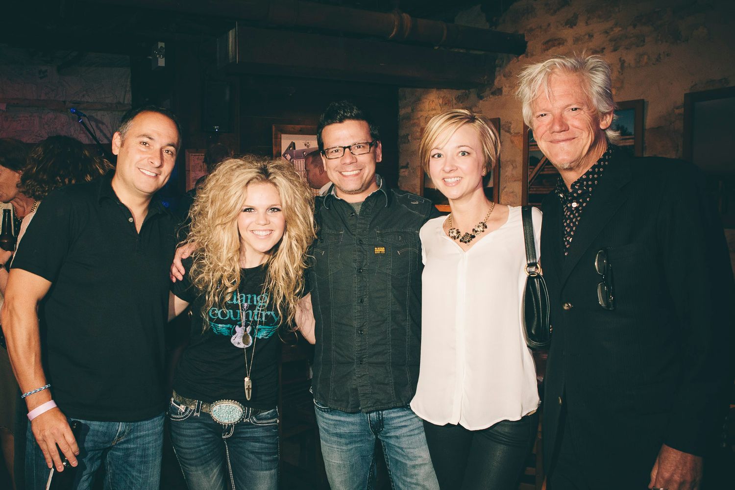 Five people smile together for a group photo at an indoor country music event.