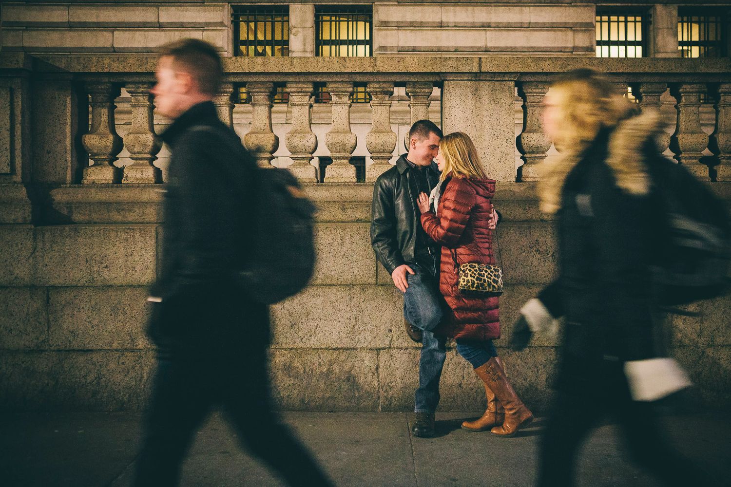Couple sharing intimate moment against stone wall while pedestrians walk by.