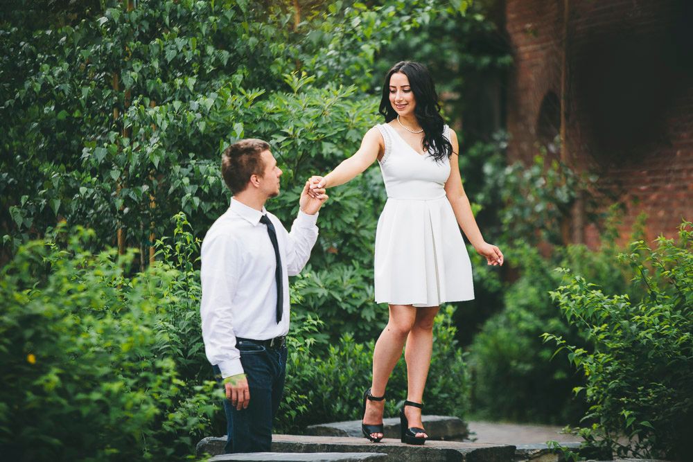 Man kneeling on one knee holding woman's hand in a lush green garden setting.