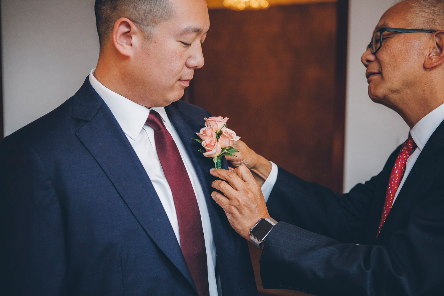 Older man pinning a pink floral boutonniere on groom in navy suit with burgundy tie.