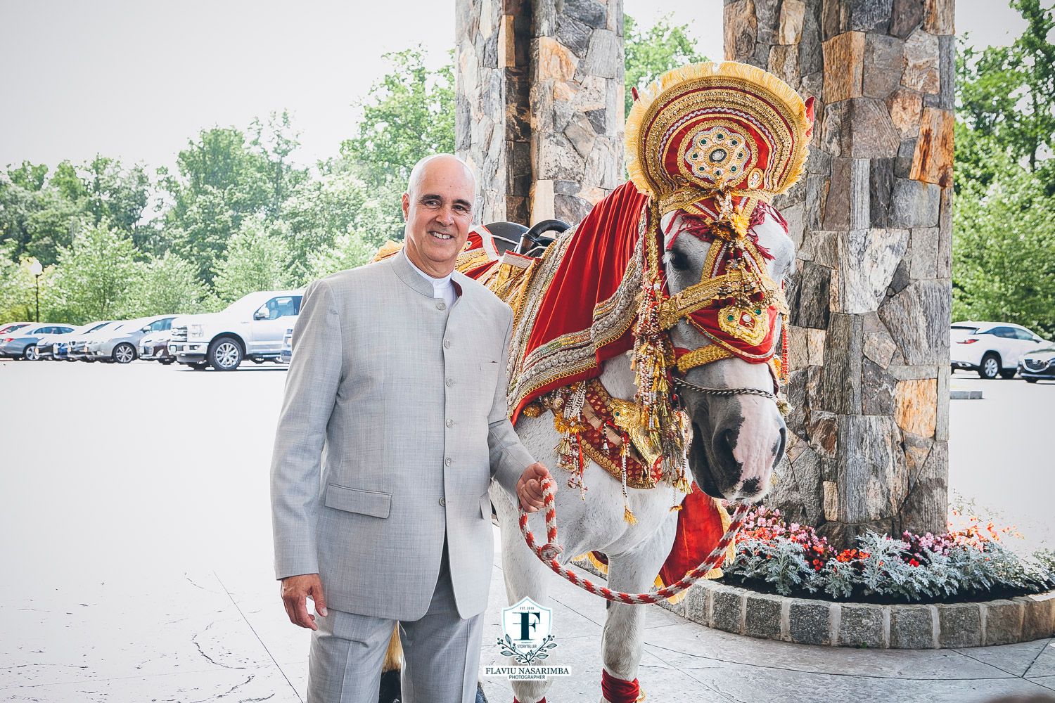 Groom in grey suit poses with colorful traditional Kathakali dancer outside stone wedding venue.