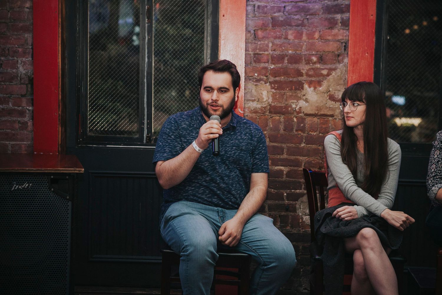 Man speaking into microphone while seated on stool in front of brick wall at panel event.