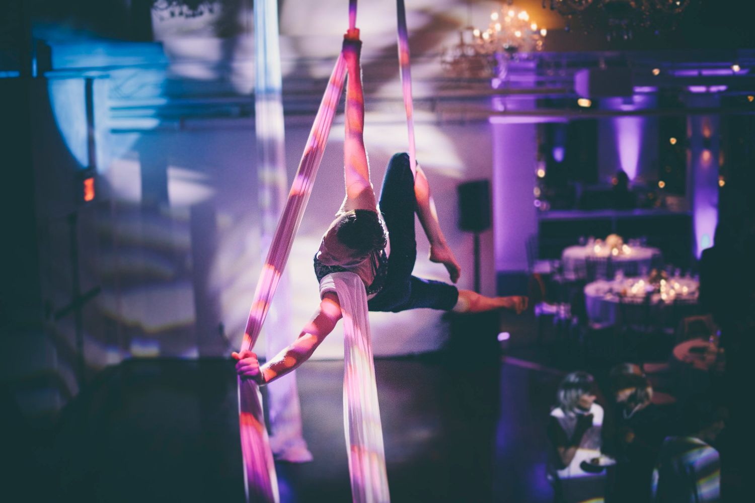 Aerial silk performer gracefully suspended in pink ribbons during an elegant event with purple lighting.