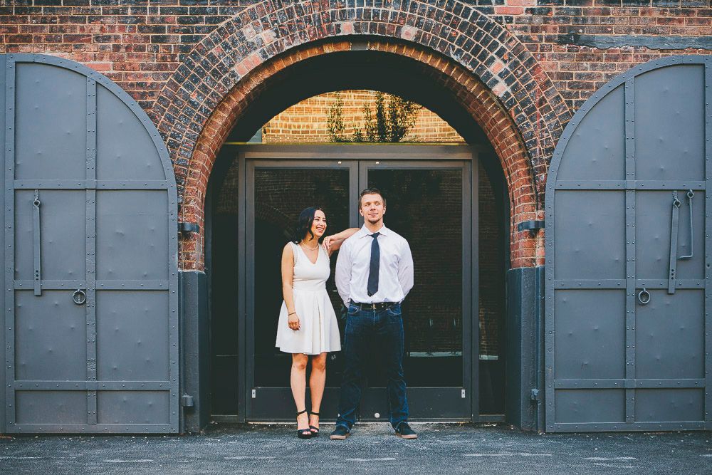 Couple posing together in front of brick archway with grey doors, woman in white dress and man in white shirt with tie.