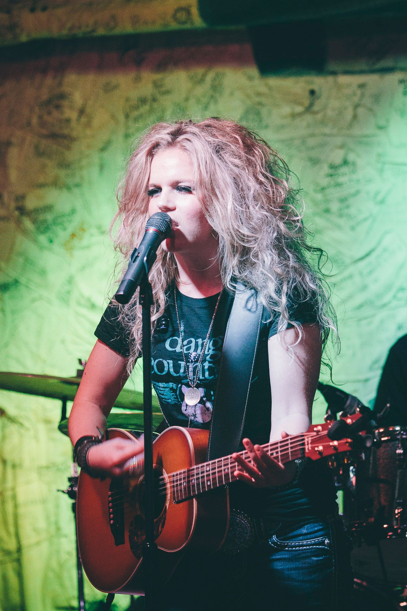 Curly-haired blonde female singer performs acoustic guitar on stage under green stage lighting.