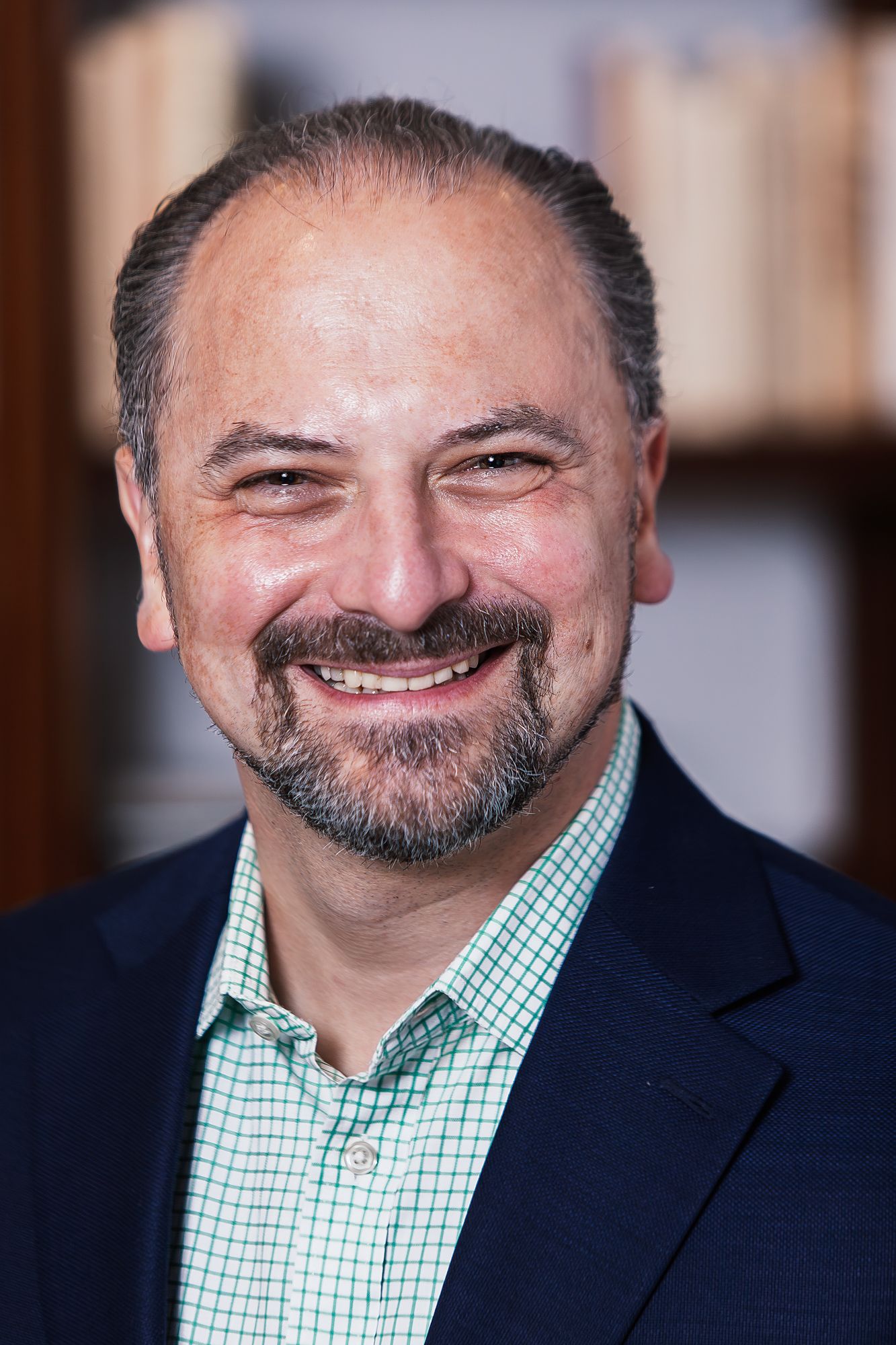 Professional headshot of smiling bearded man in navy blazer and checkered shirt in front of bookshelf.