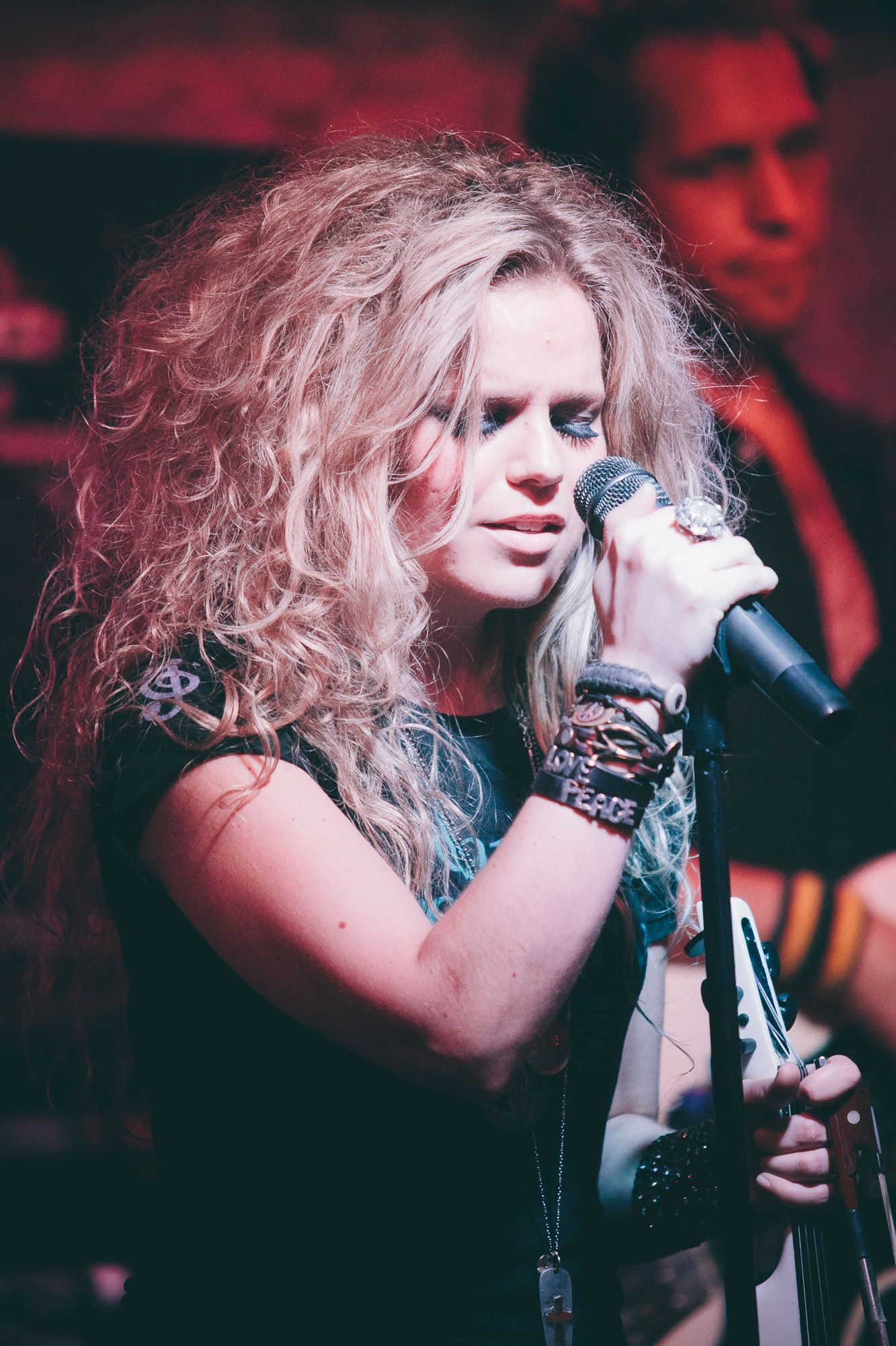 Curly blonde female vocalist sings passionately into microphone during live stage performance.