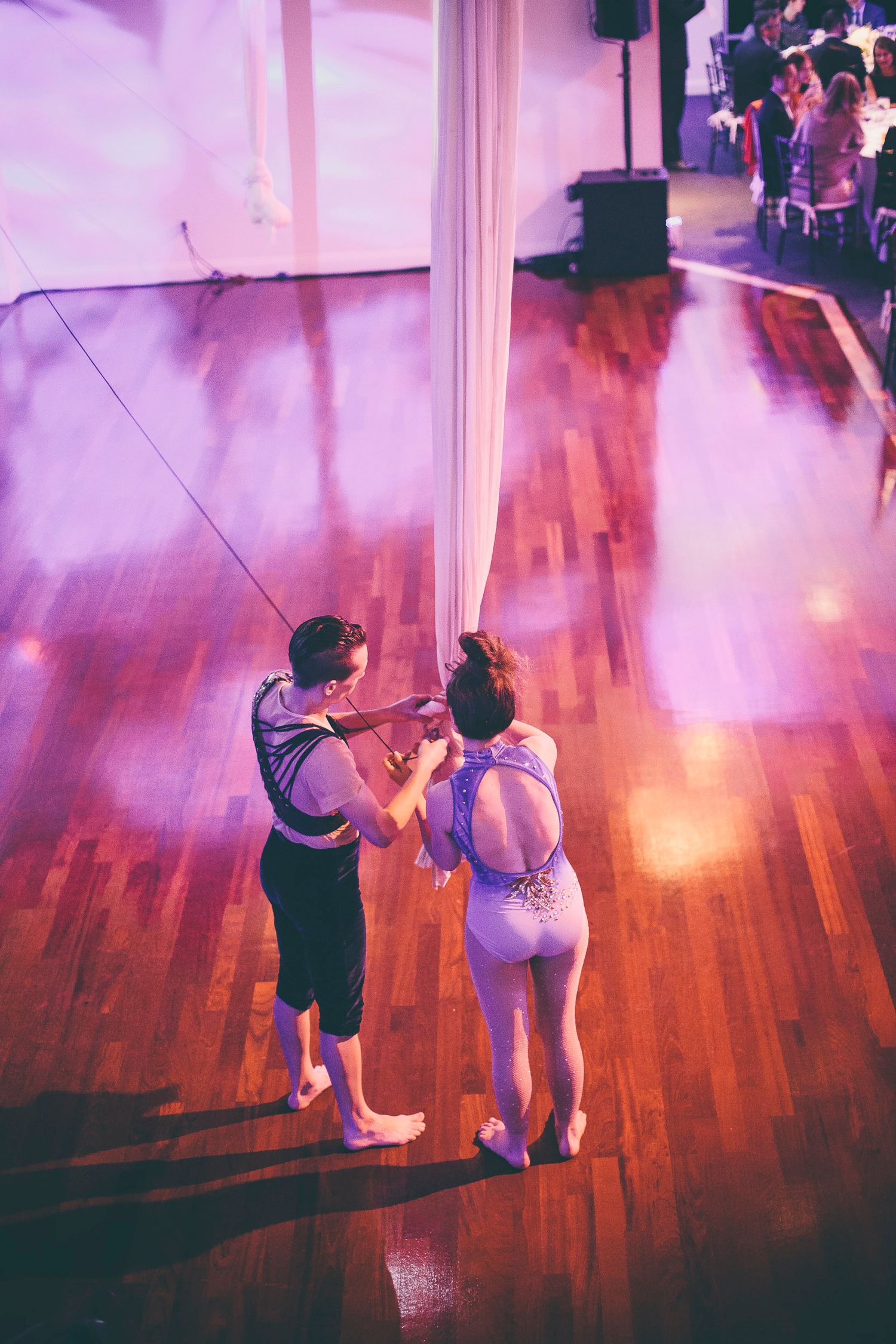 A violinist performs with an aerial silk dancer on a hardwood floor under purple stage lighting.