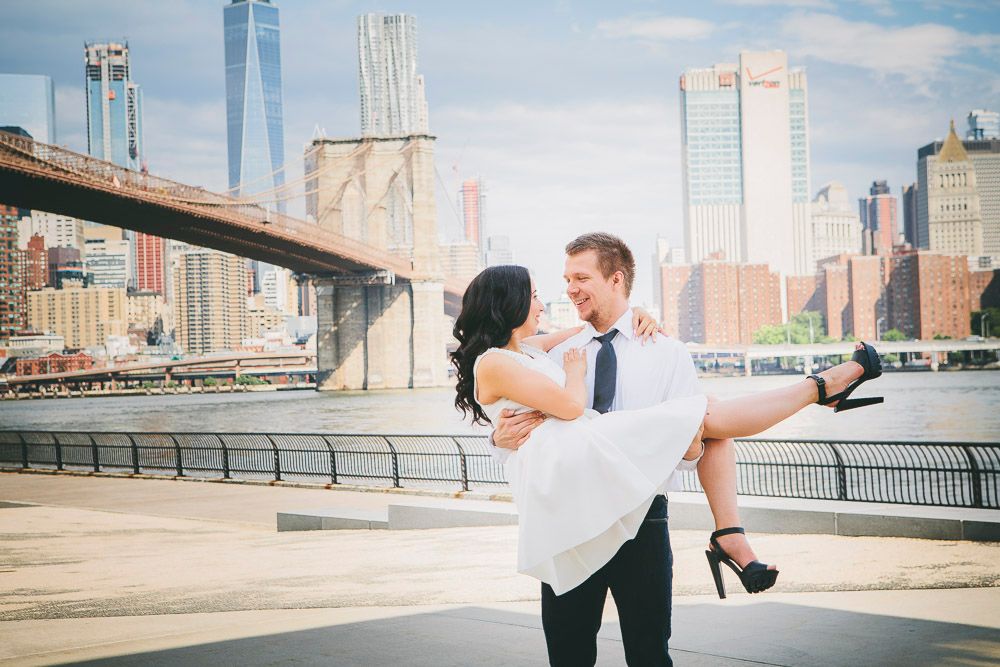 Man lifting woman in white dress near Brooklyn Bridge with New York City skyline in sunny background.