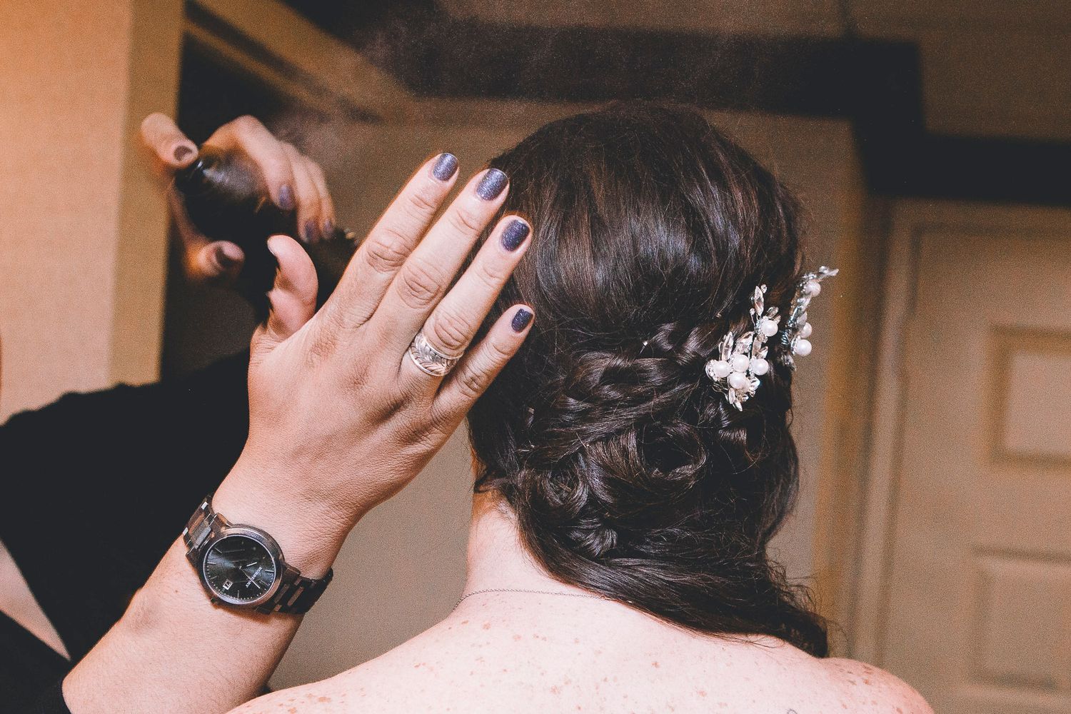 Hairstylist spraying a bride's elegant dark updo adorned with delicate floral hair accessories before wedding.