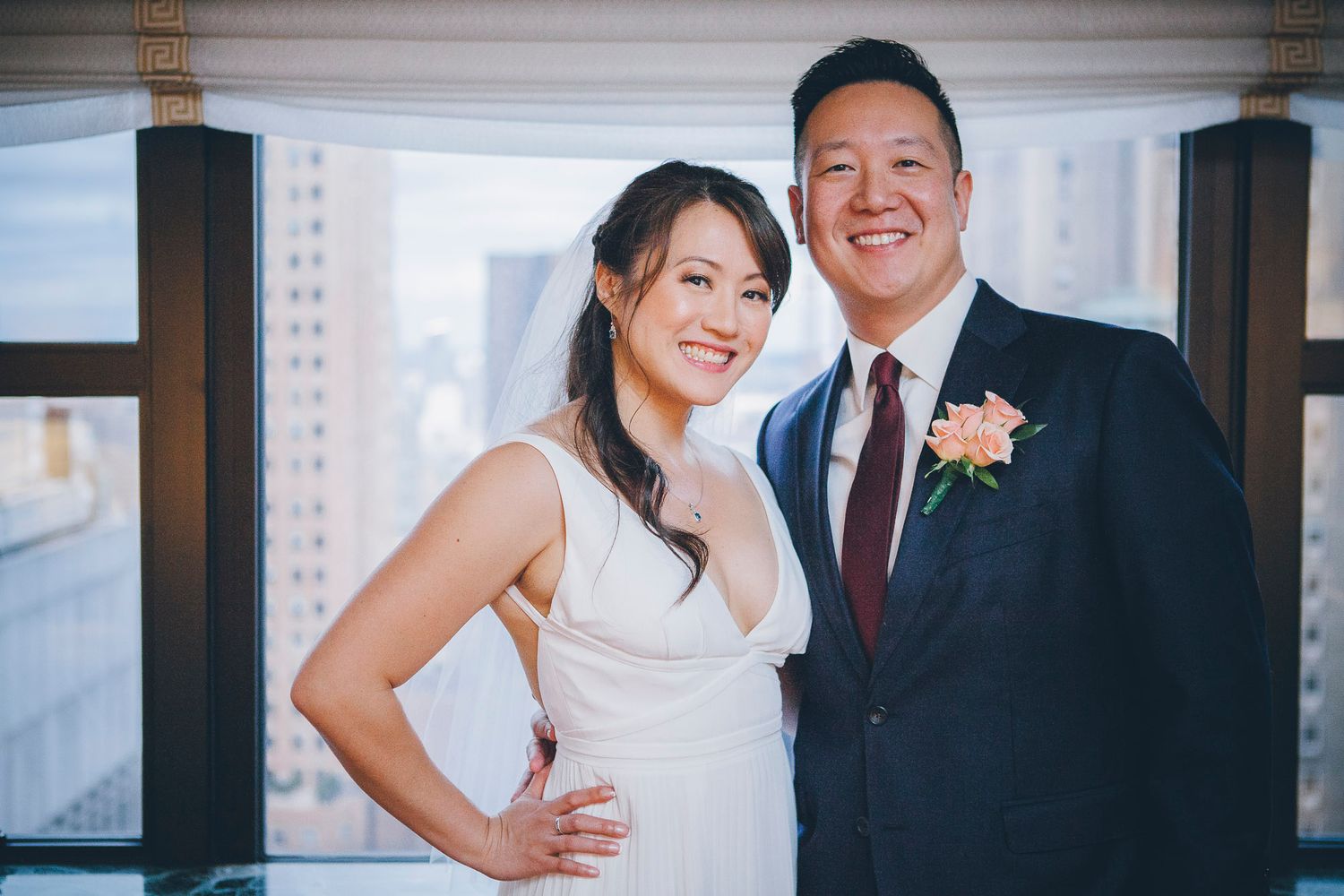 Happy bride and groom pose together near a window with urban cityscape in the background.