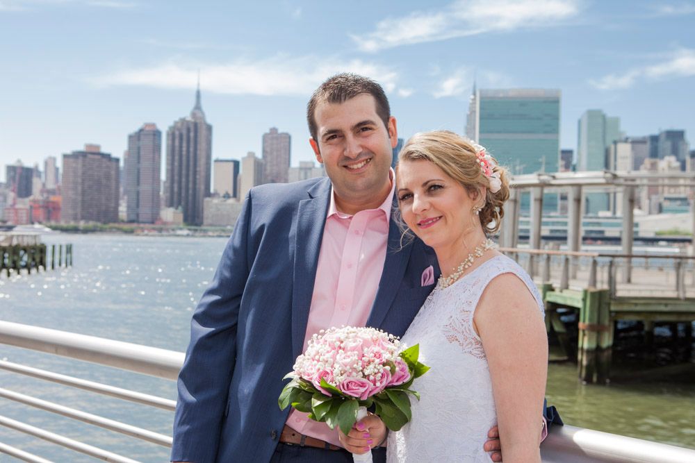 Smiling bride and groom pose with pink bouquet against New York City skyline waterfront.