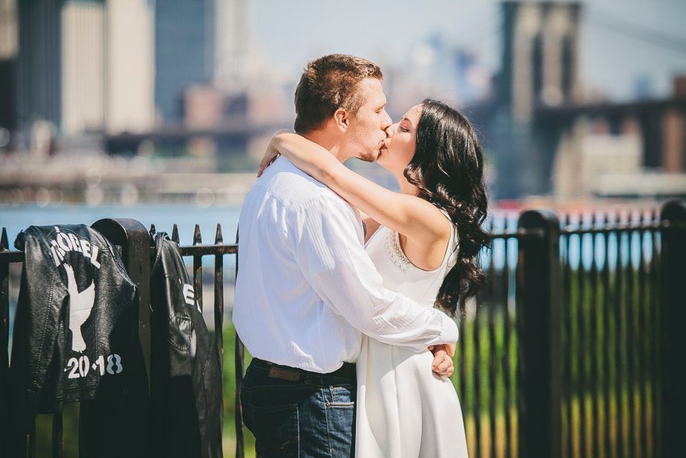 Couple sharing a romantic kiss near Brooklyn Bridge with NYC skyline behind them.