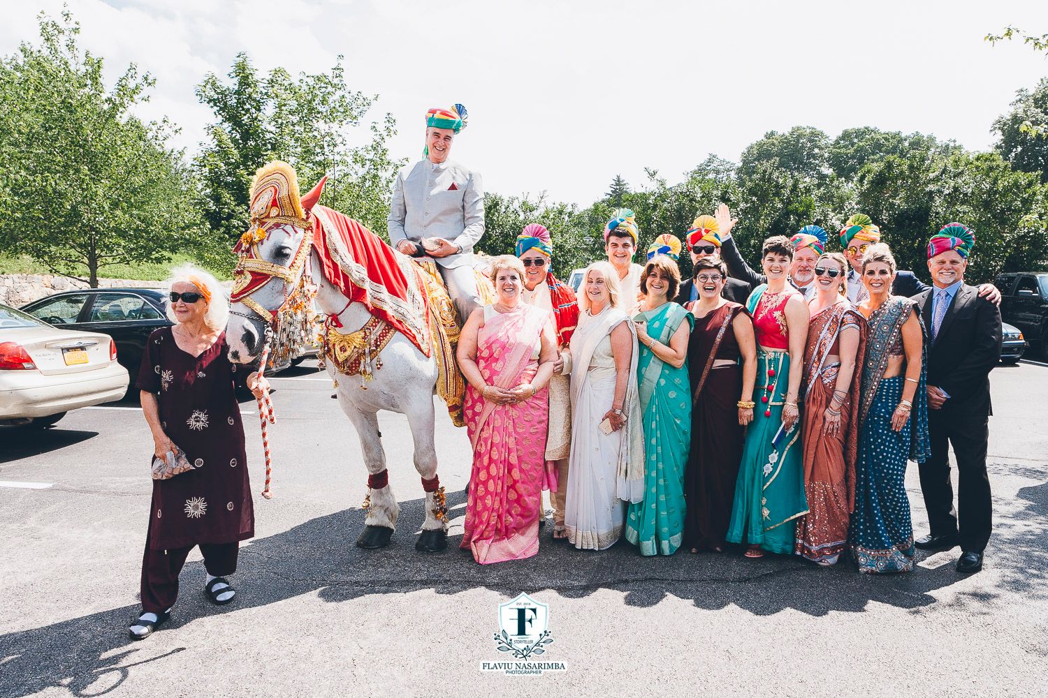Wedding party in colorful Indian attire poses with decorated white horse during baraat procession.