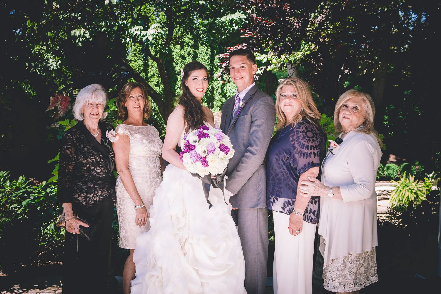 Bride and groom pose with four female family members holding purple bouquet in garden setting.