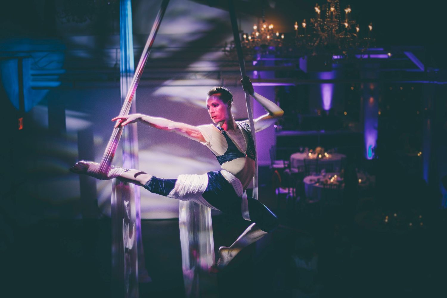 Female aerialist in costume performing a split on aerial silks in a chandelier-lit ballroom.