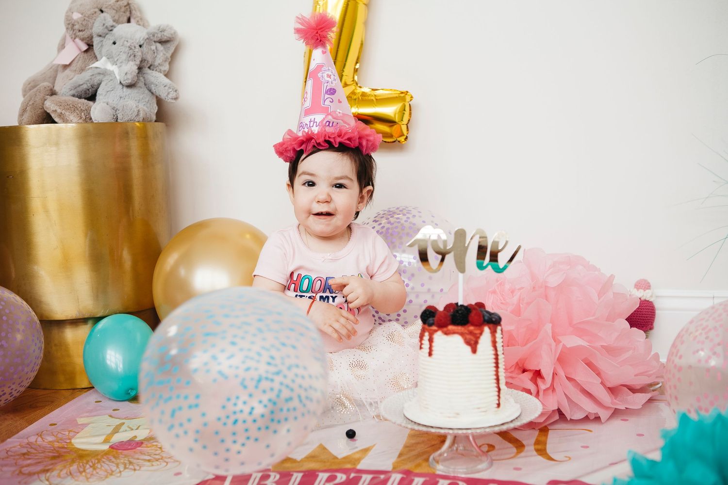 Baby girl in pink birthday hat sits beside a decorated first birthday cake surrounded by festive balloons.