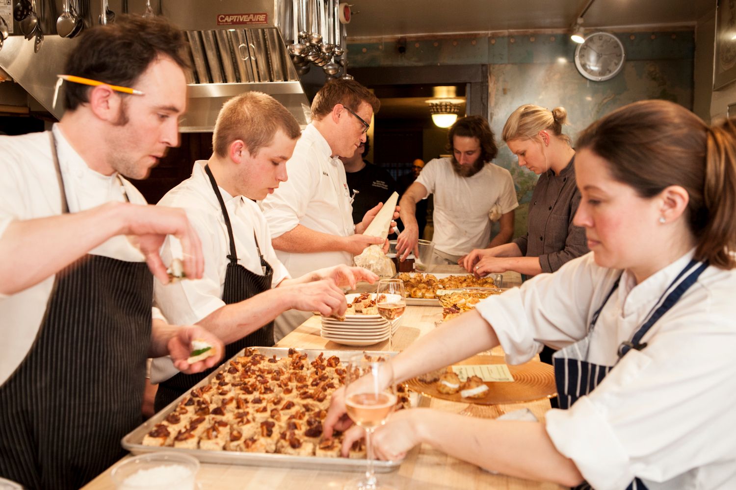 Team of chefs plating bite-sized hors d'oeuvres on trays in a busy restaurant kitchen.