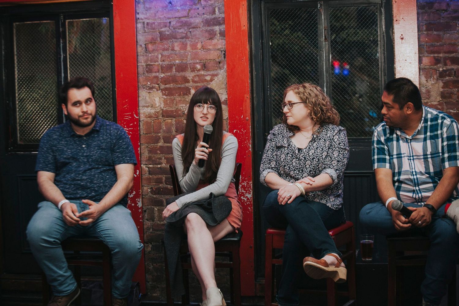Four panelists seated on stools during a discussion event in front of a red brick wall.