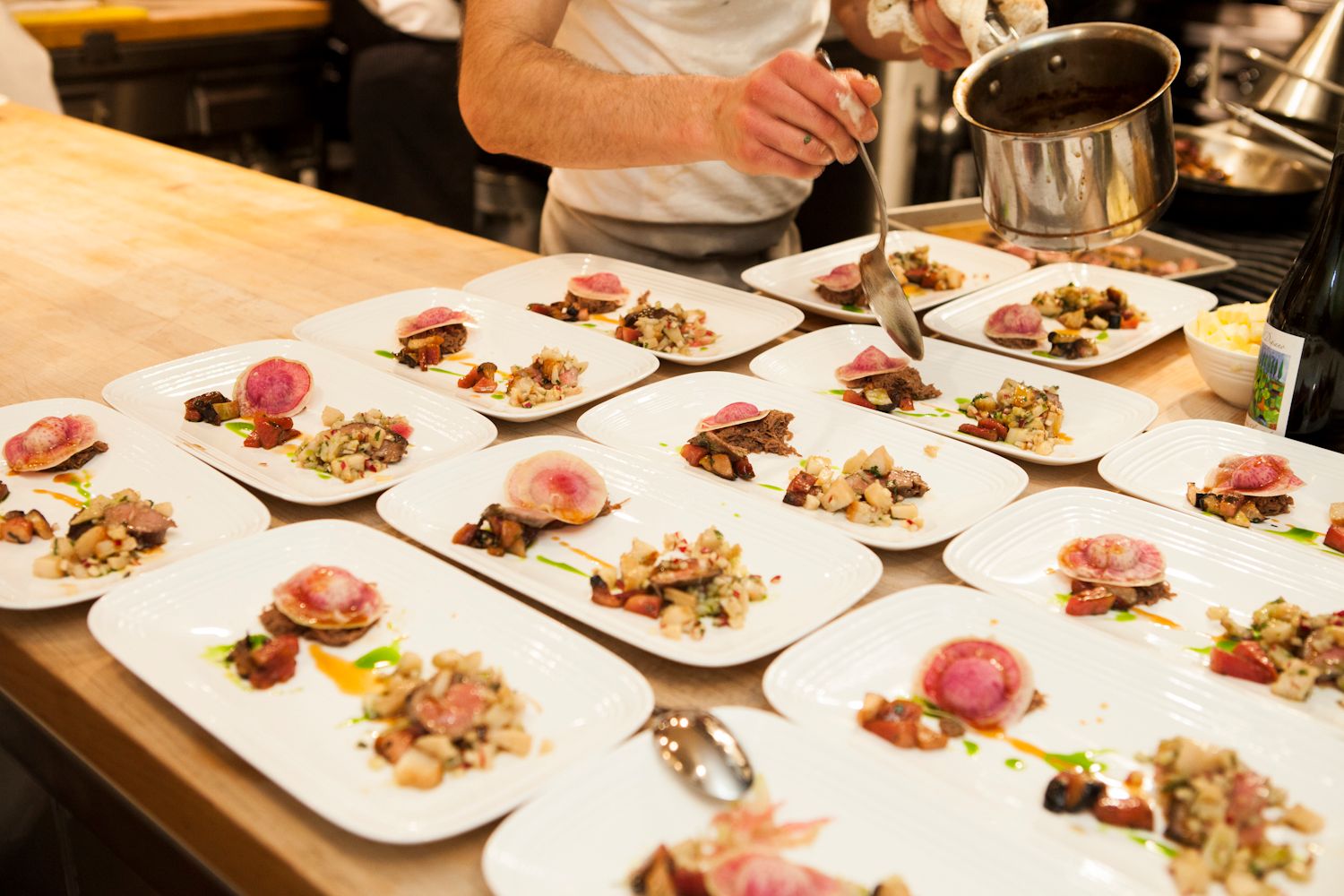 Chef plating multiple identical gourmet dishes with seared meat and colorful garnishes for service.