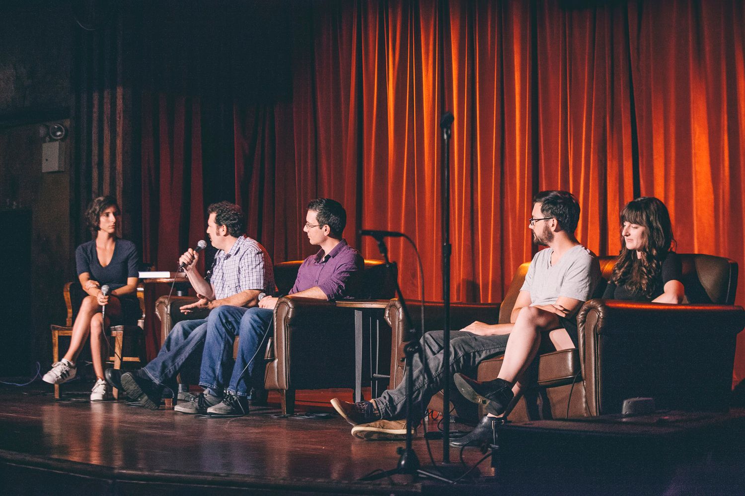 Five panelists seated in chairs on stage during panel discussion with red curtain backdrop.