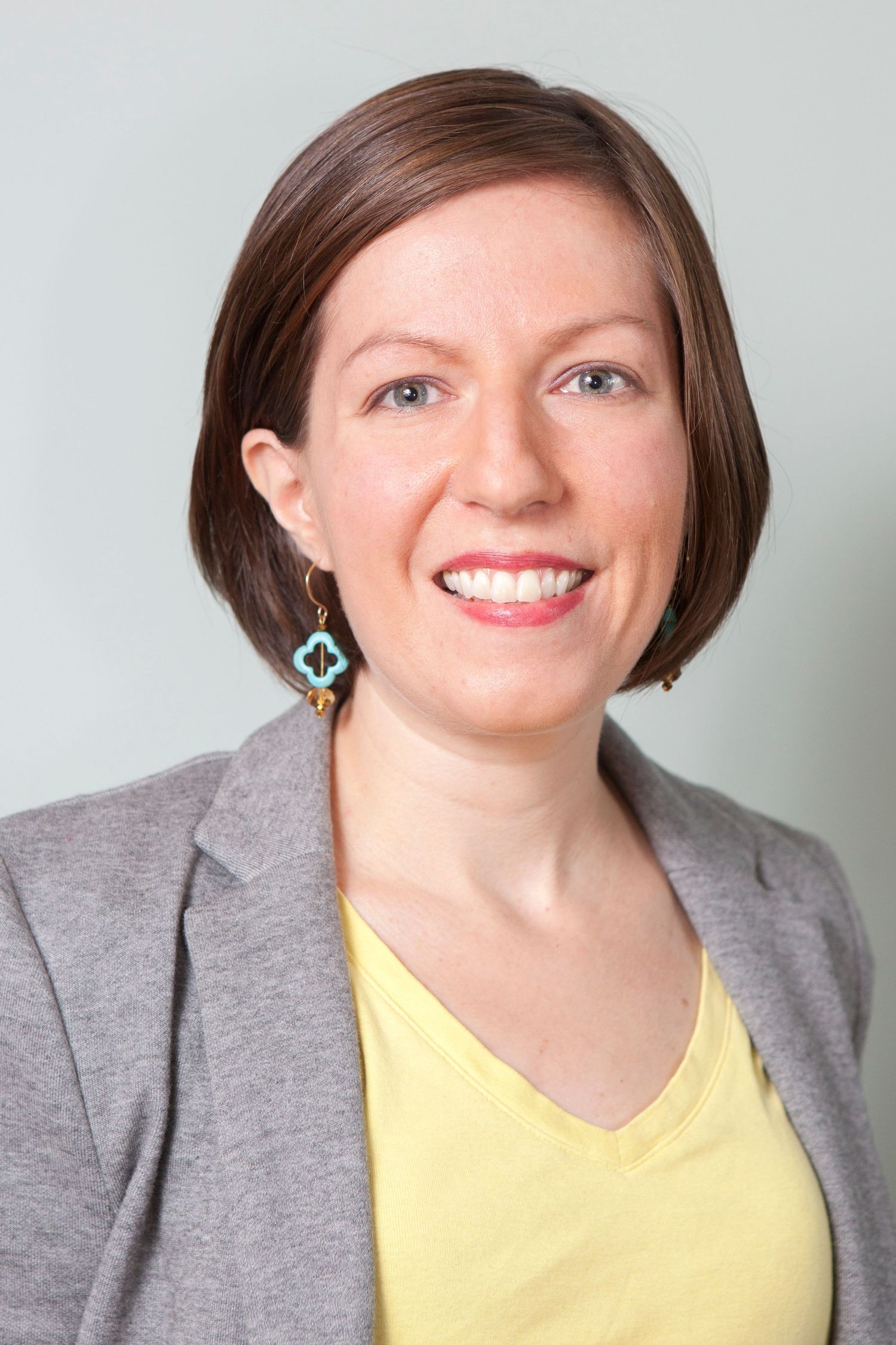 Smiling brunette woman in a gray blazer and yellow top wearing turquoise earrings, headshot.