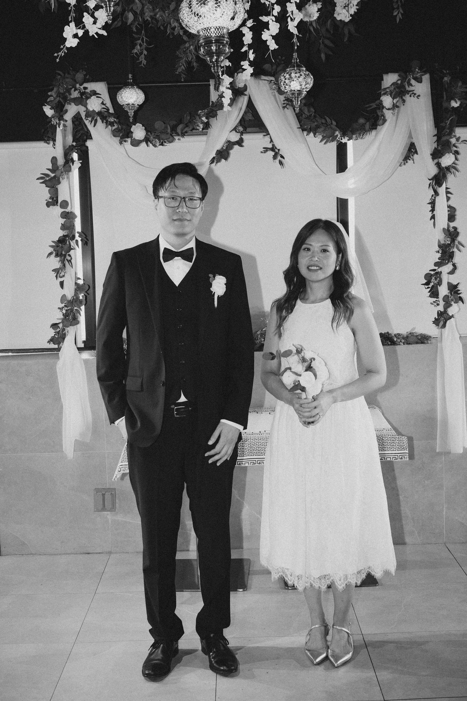 Groom in black tuxedo and bride in white dress stand together under decorated wedding arch.
