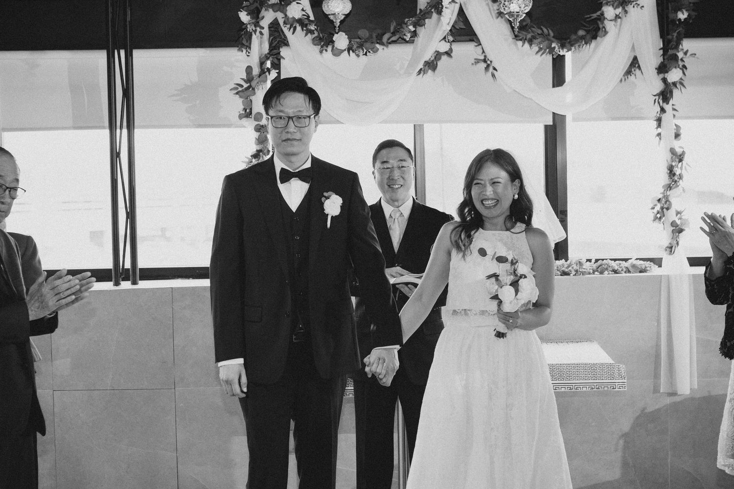 Newlywed Asian couple holds hands and smiles after wedding ceremony as guests applaud.