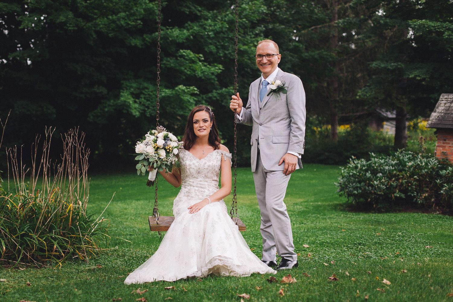 Bride sitting on a garden swing holding a bouquet while groom stands beside her on green grass.
