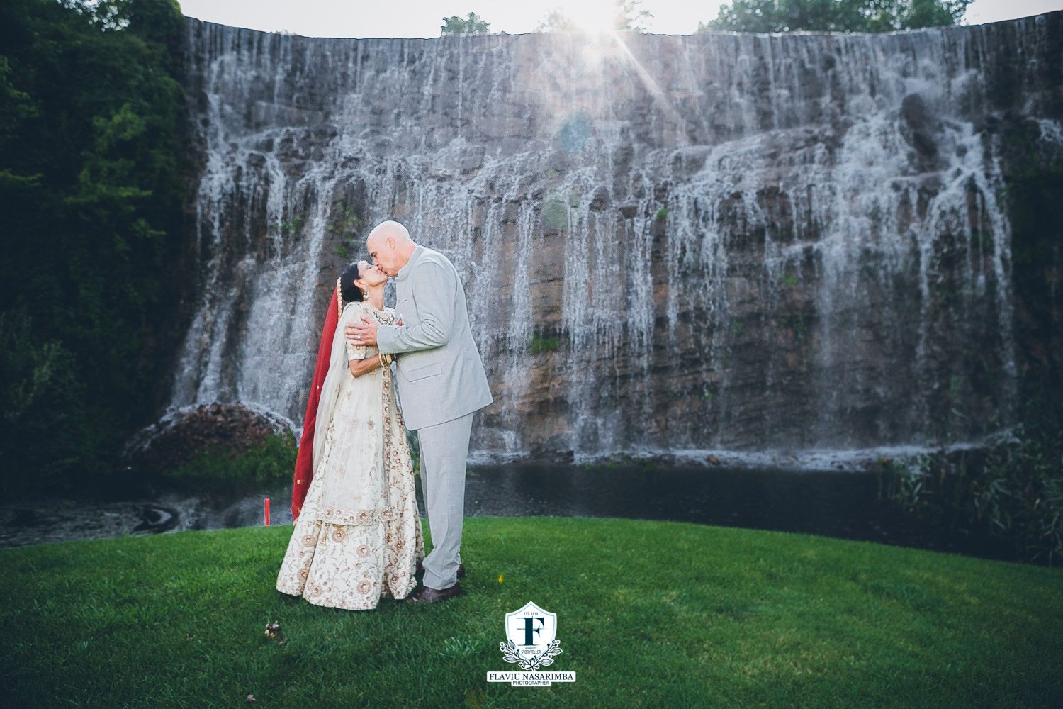Bride and groom share romantic kiss in front of stunning waterfall backdrop during Indian wedding ceremony outdoors.
