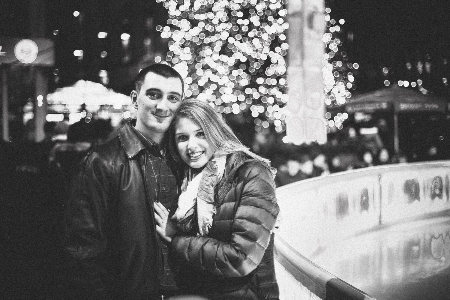 Black and white photo of smiling couple embracing near Christmas tree at night.