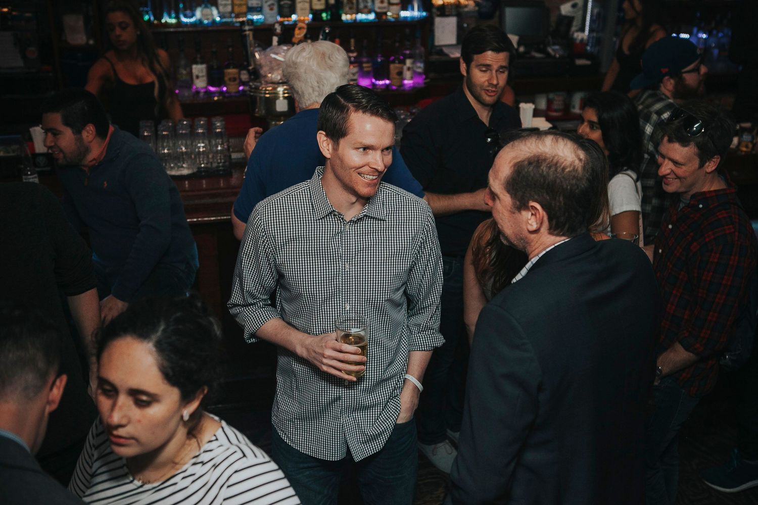 Man in gray shirt holds a drink while chatting at a crowded bar event.