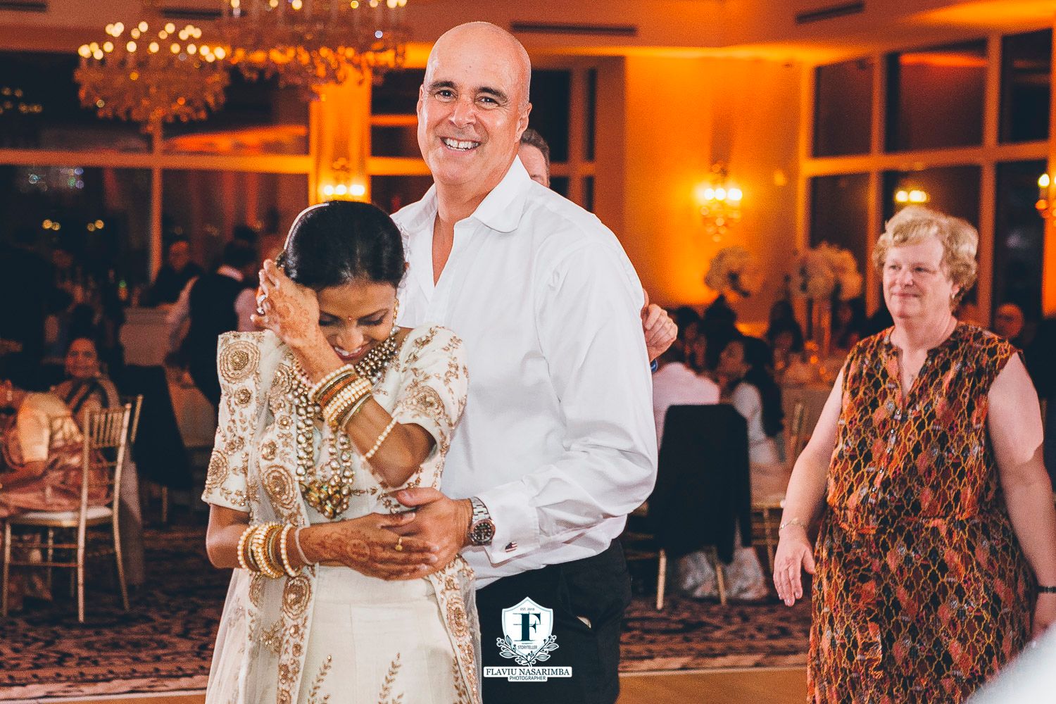 Indian bride in white lehenga dances joyfully with groom in white shirt at warm-lit wedding reception.
