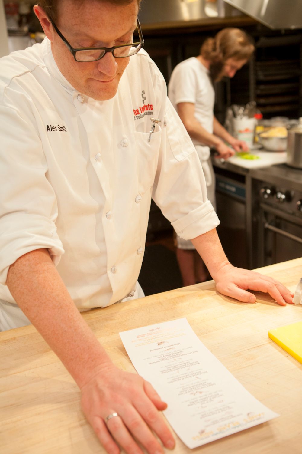 Chef in white uniform reviewing a printed menu on a wooden prep counter in a kitchen.