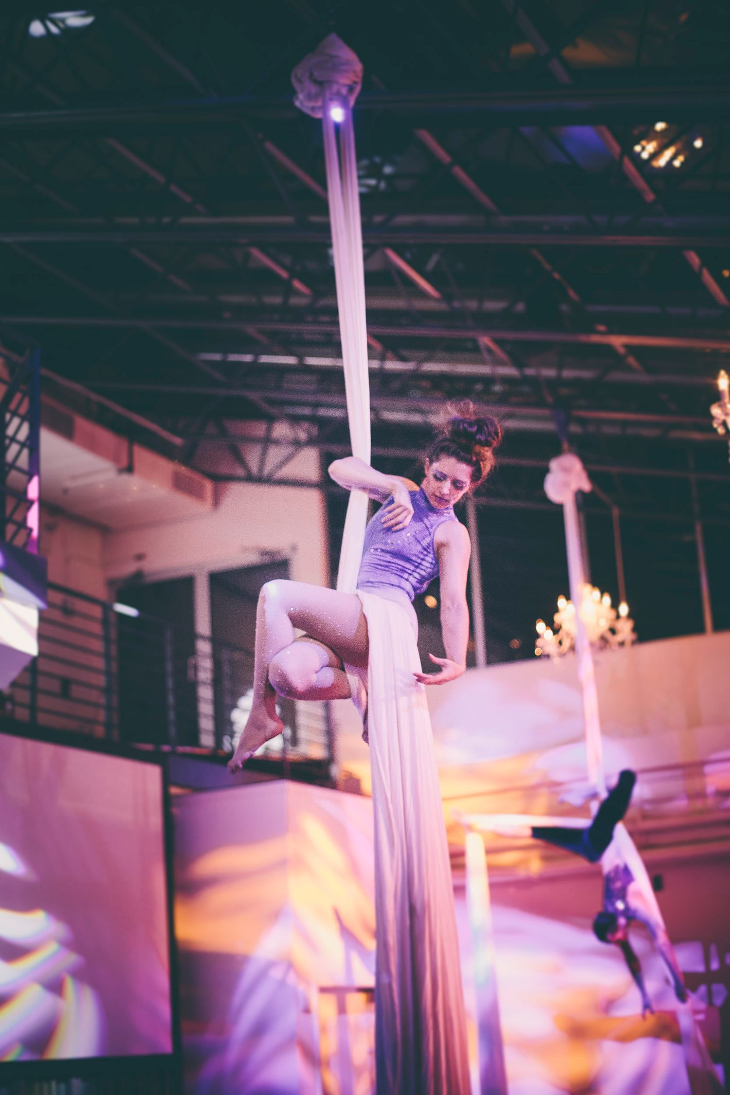 A smiling aerial silk performer poses gracefully on white fabric in a purple-lit event venue.