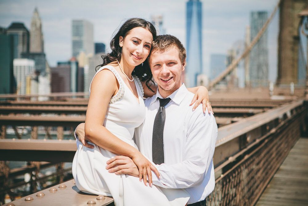 Smiling couple embracing on Brooklyn Bridge with One World Trade Center and NYC skyline visible behind them.