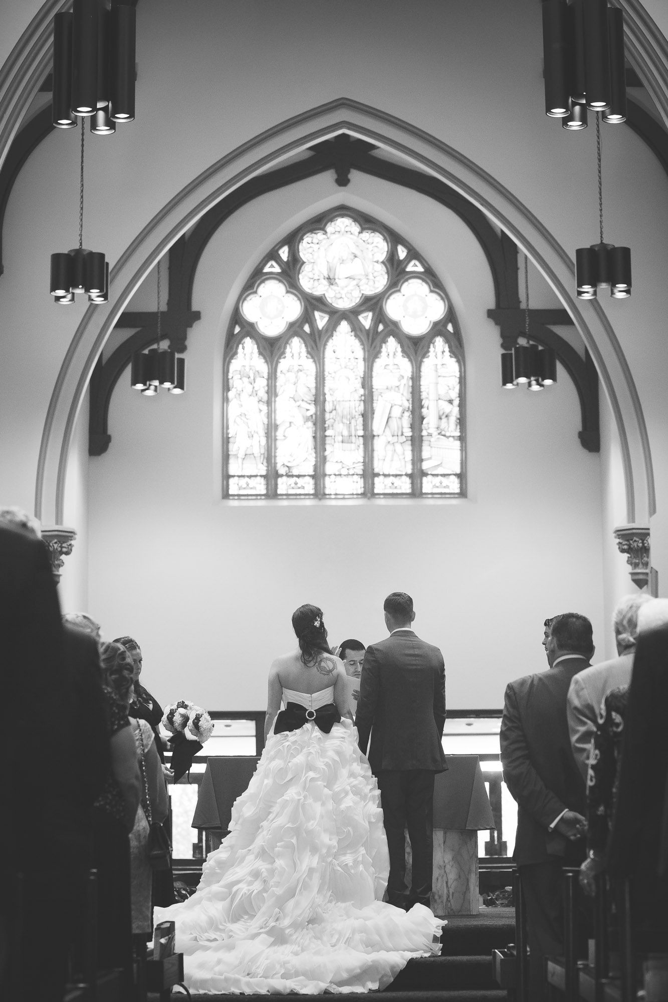 Bride and groom stand at church altar beneath gothic stained glass window during wedding ceremony.