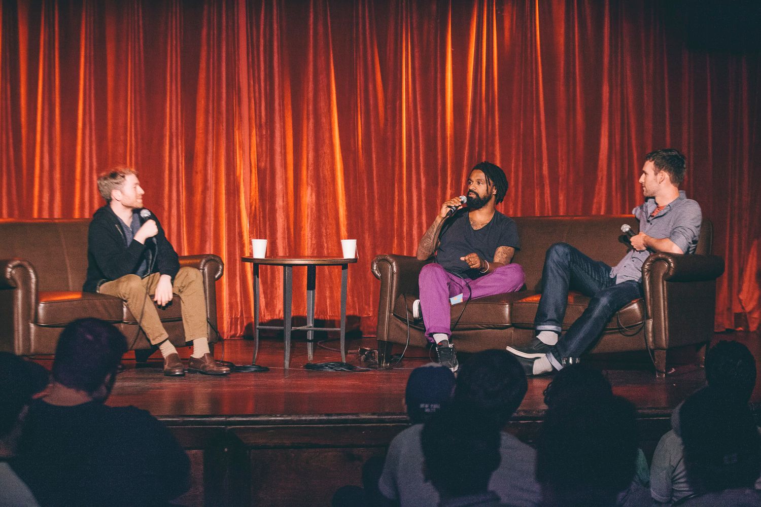Three men having a conversation on stage at a comedy club with red curtain backdrop.
