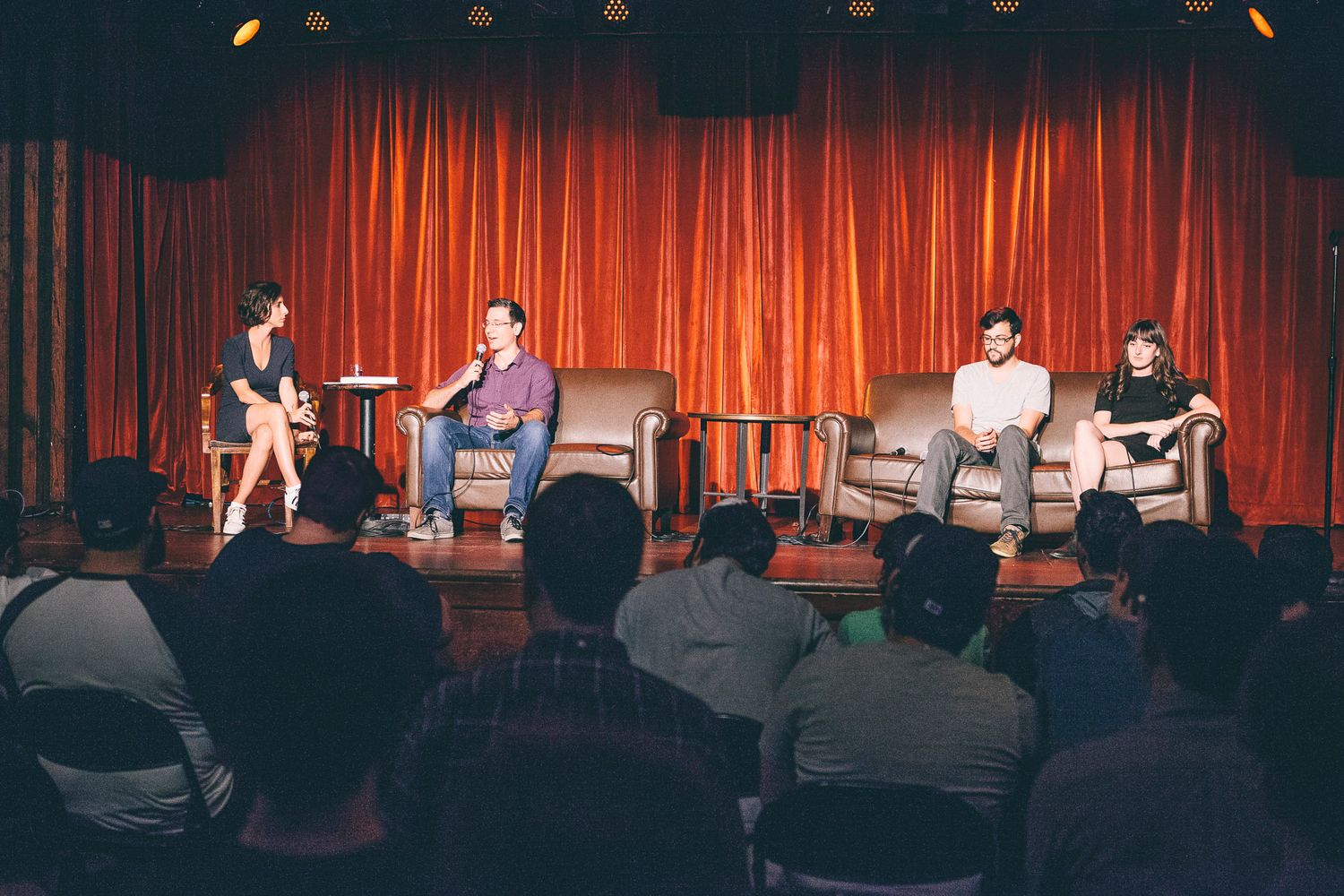 Four panelists seated in chairs on a stage with orange curtain backdrop at a live event.